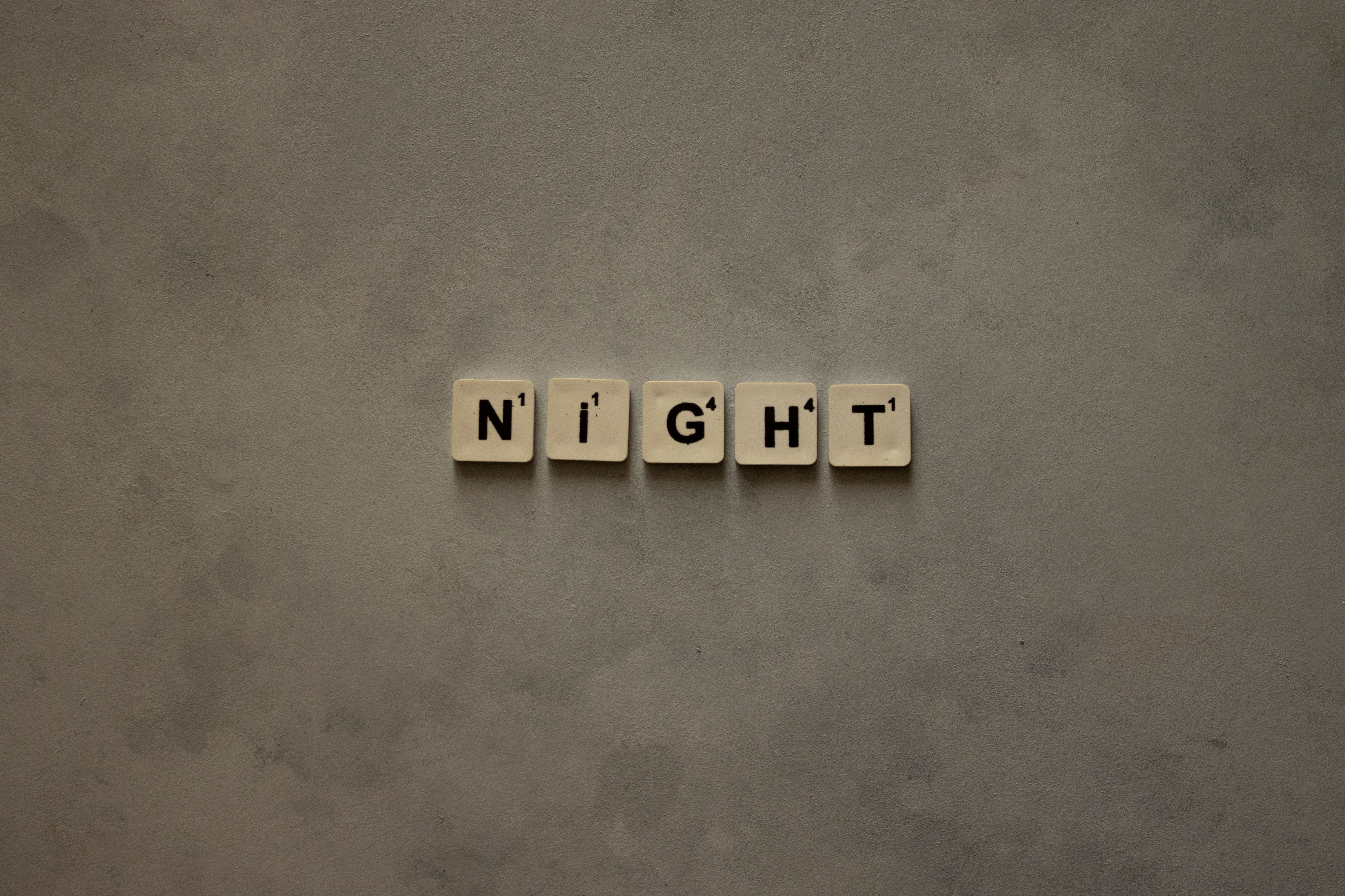 Scrabble tiles arranged to spell 'Night' on a minimalist gray background.
