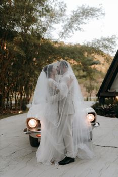 Bride and groom share a romantic kiss outdoors in front of a vintage car.