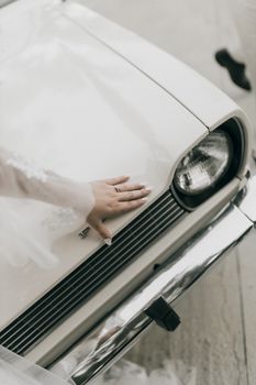 A bride's hand gracefully resting on a vintage white car's hood, creating a timeless wedding moment.