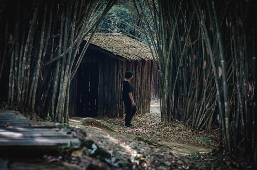 A man standing near a rustic bamboo hut in a dense forest path in Thailand.