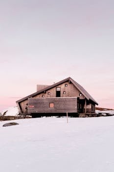 A wooden cabin stands amidst snow, under a soft pink sky, capturing winter serenity.