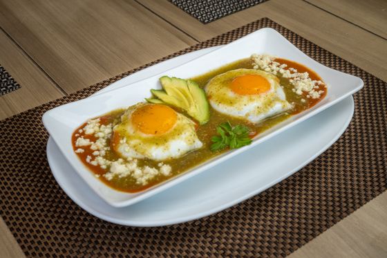 Two sunny-side-up eggs in green salsa with red sauce, avocado slices and crumbled cheese