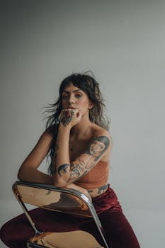 Stylish tattooed woman sitting on a chair, with trendy fashion and thoughtful expression.