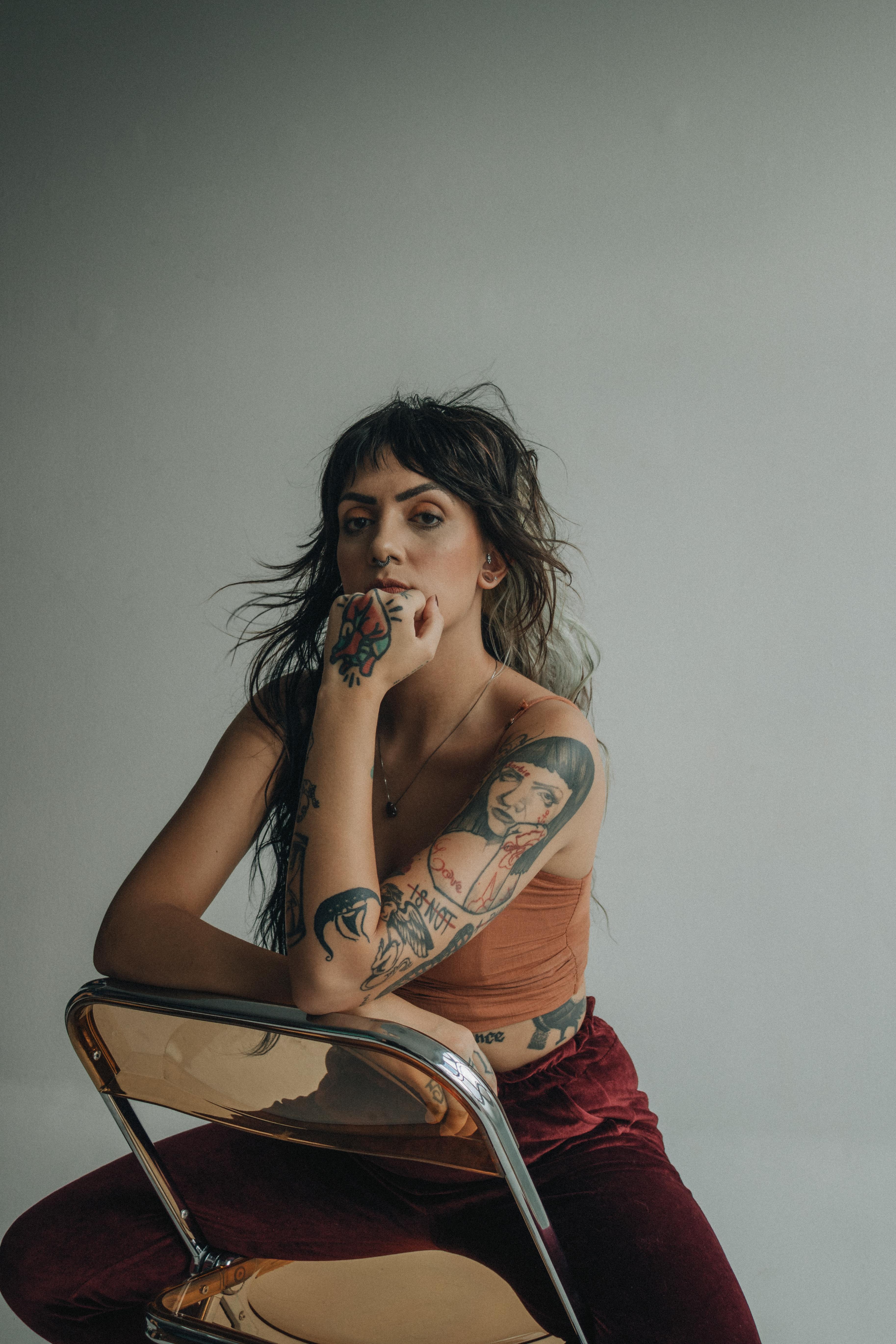 Gratis Mujer tatuada con estilo sentada en una silla, con moda actual y expresión pensativa. Foto de stock