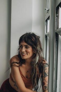 A woman with tattoos smiles while leaning by a window indoors, exuding confidence and style.