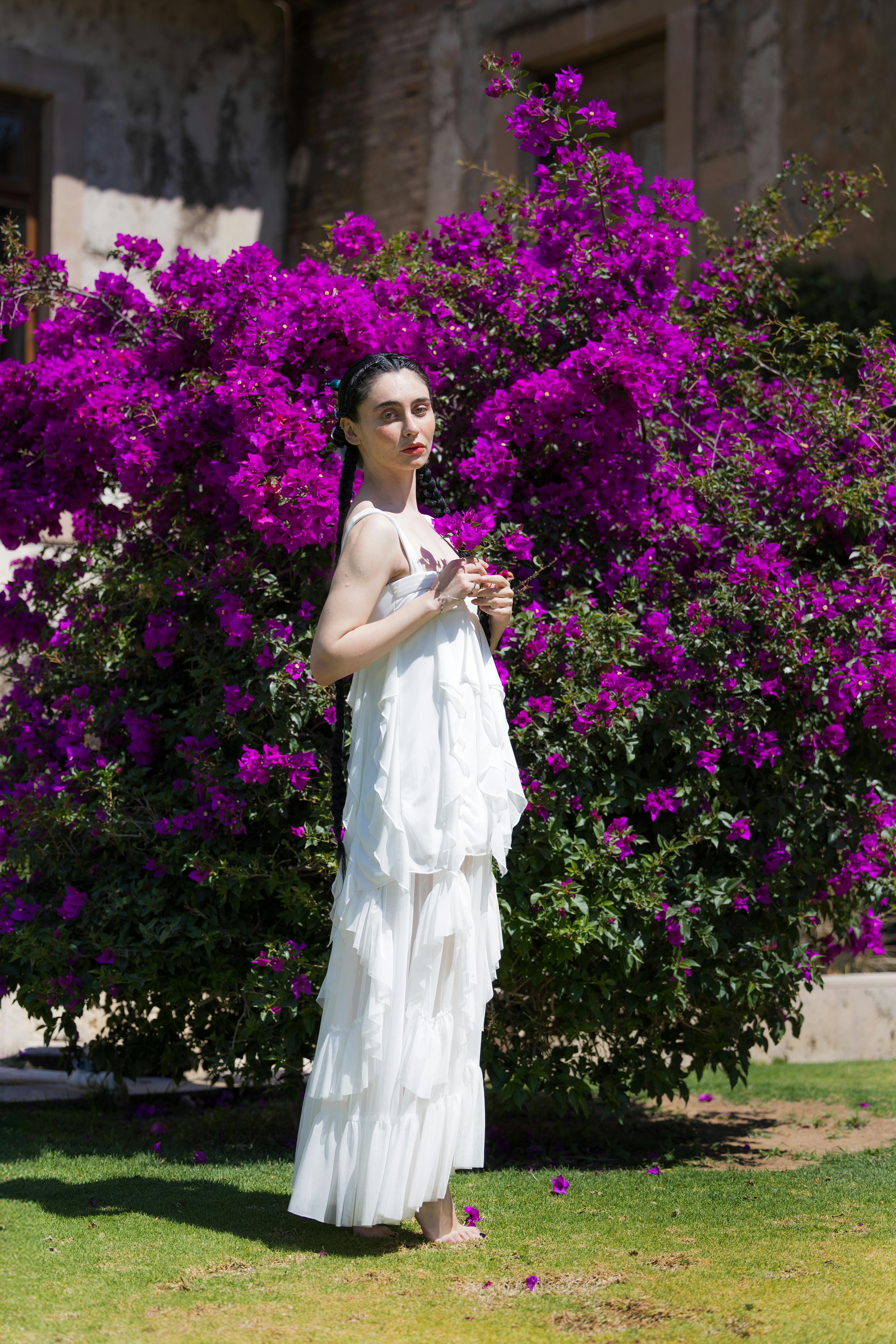 De franc Una dona amb vestit blanc es troba al costat d'una buguenvíl·lea morada vibrant en un jardí assolellat. Foto d'estoc