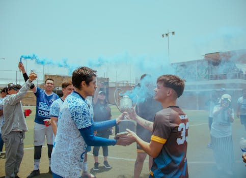 Soccer team celebrating victory with trophy, surrounded by blue smoke on outdoor field.
