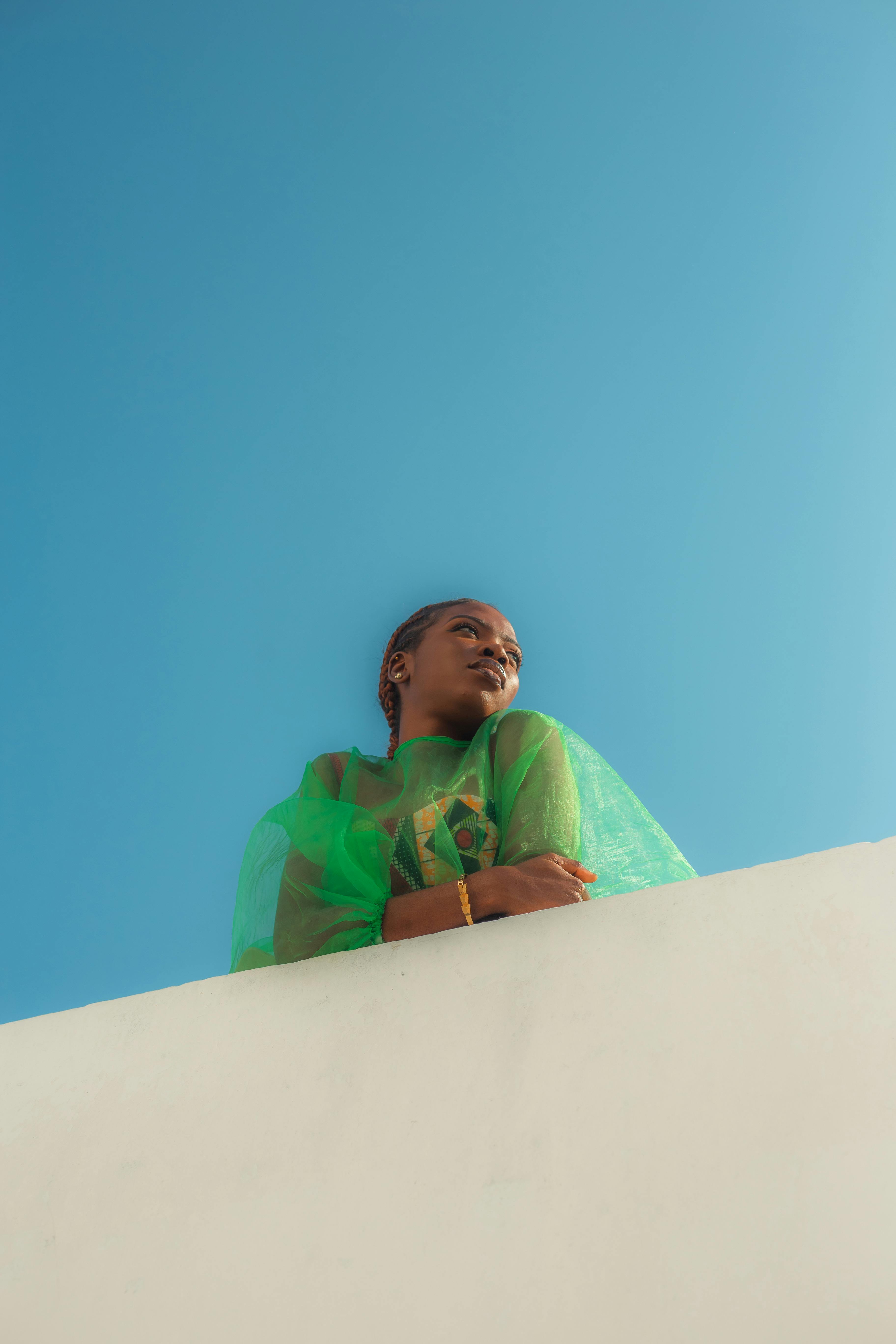 Gratuit O tânără se uită gânditoare în timp ce este îmbrăcată într-o ținută verde viu sub un cer albastru senin, surprinsă în Nigeria. Fotografie de stoc