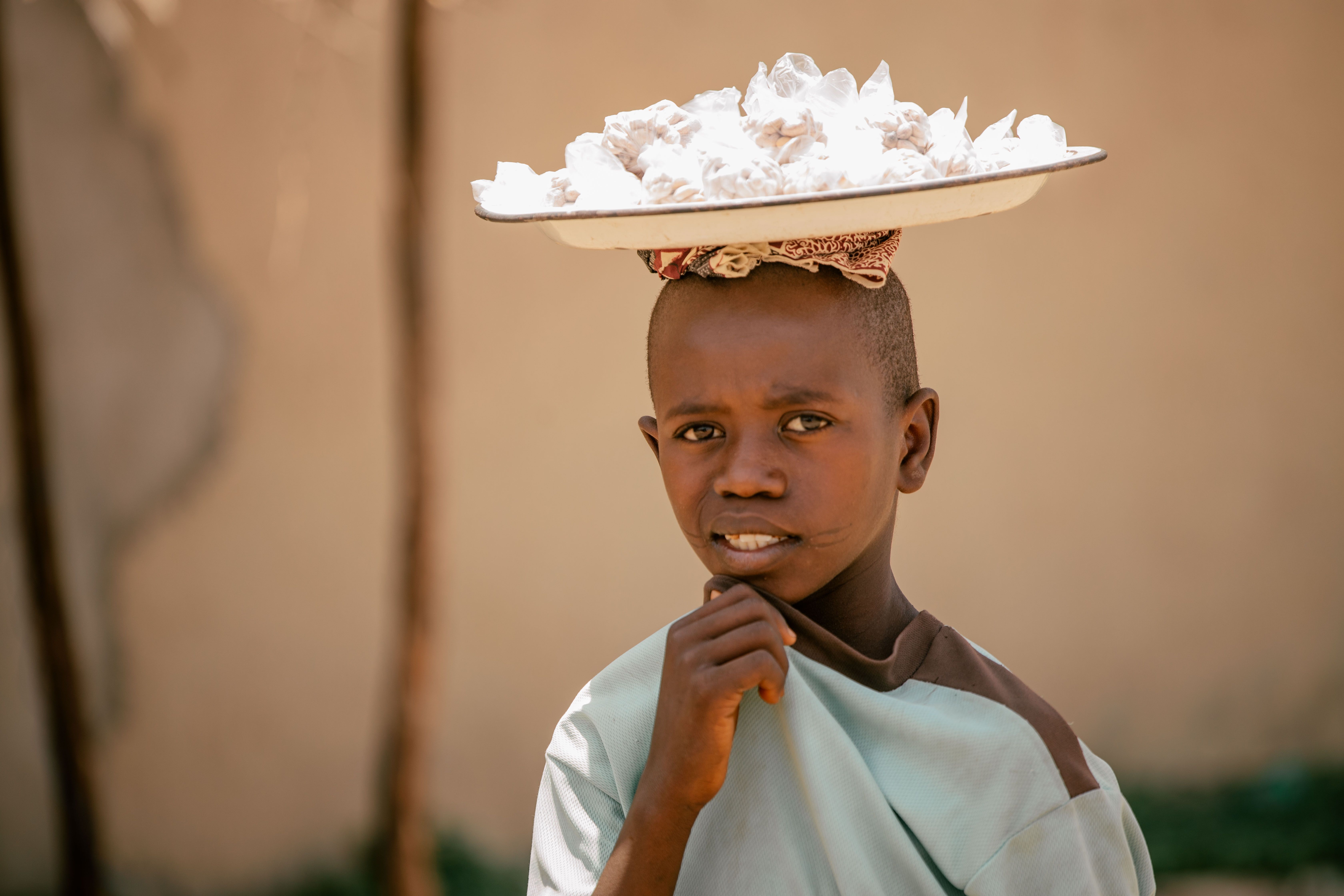 Young African boy balances a tray of white desserts on his head, showcasing skill and balance.
