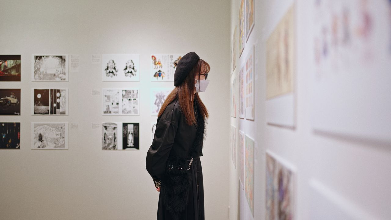 a woman wearing a mask and beret intently views art displays at a gall