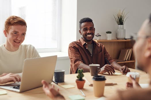 A diverse group of professionals having a casual team meeting around a wooden table in a bright, modern office.