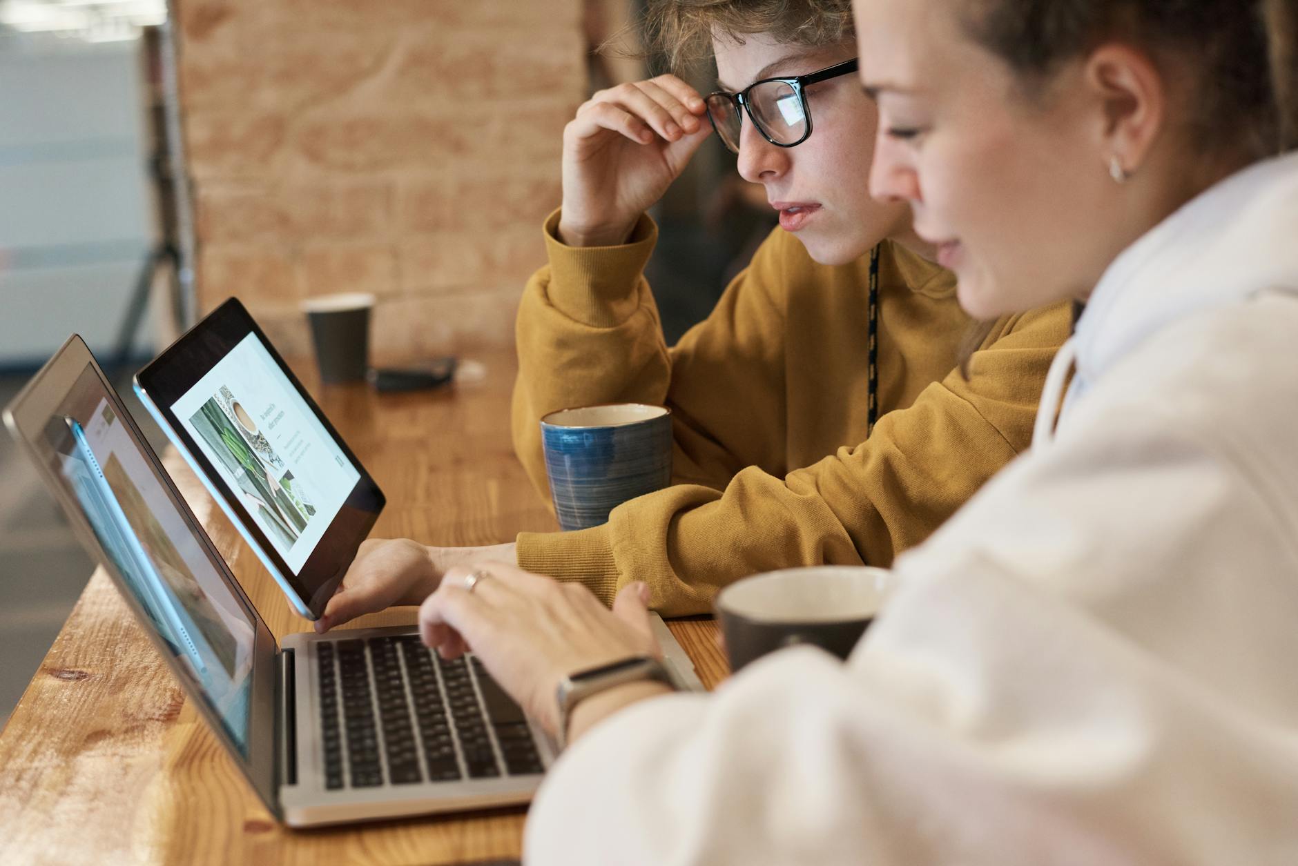 Two individuals collaborating on a project indoors, using laptops at a wooden table.