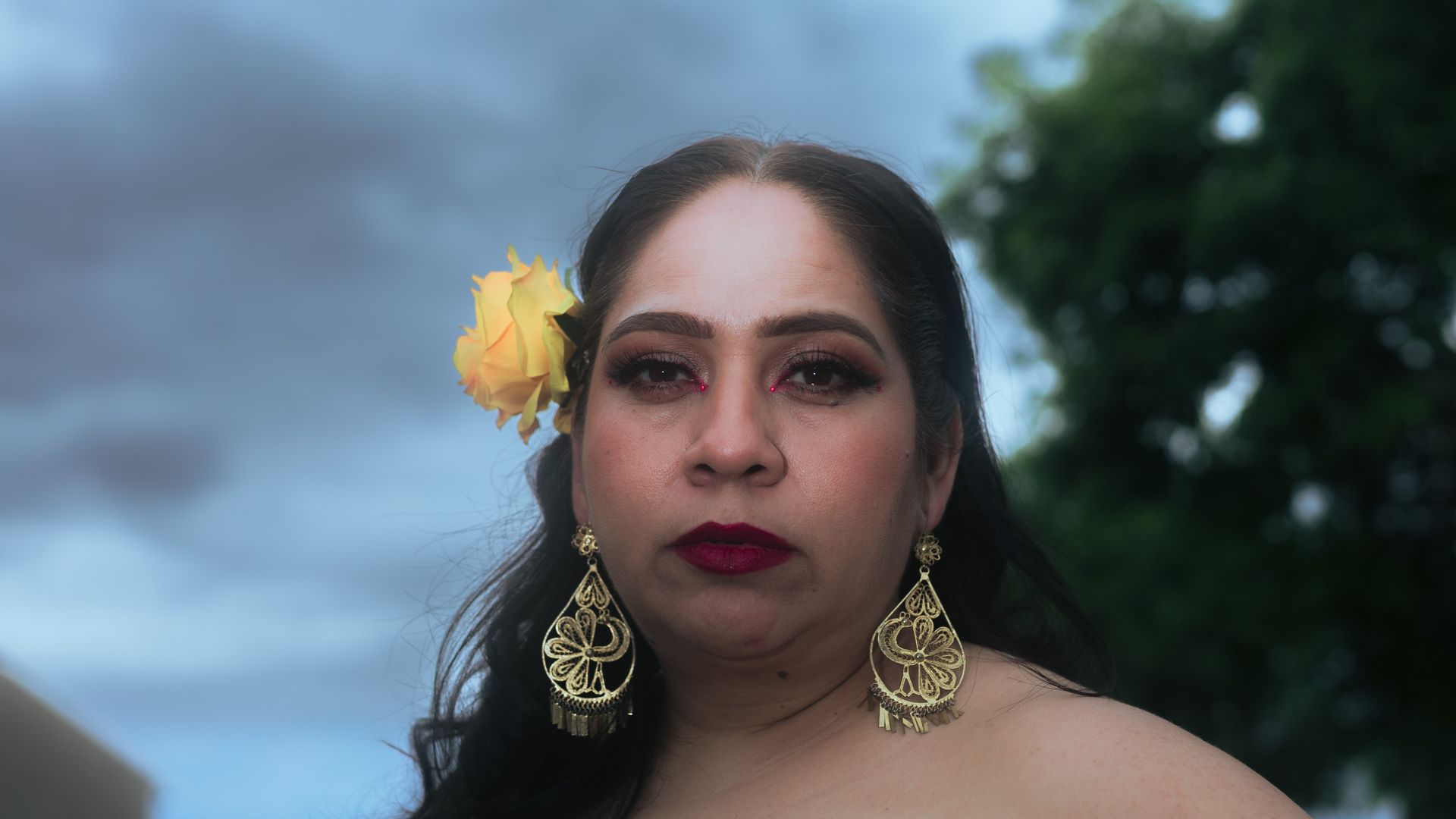 Close-up portrait of a woman with a yellow rose in her hair, featuring gold earrings.
