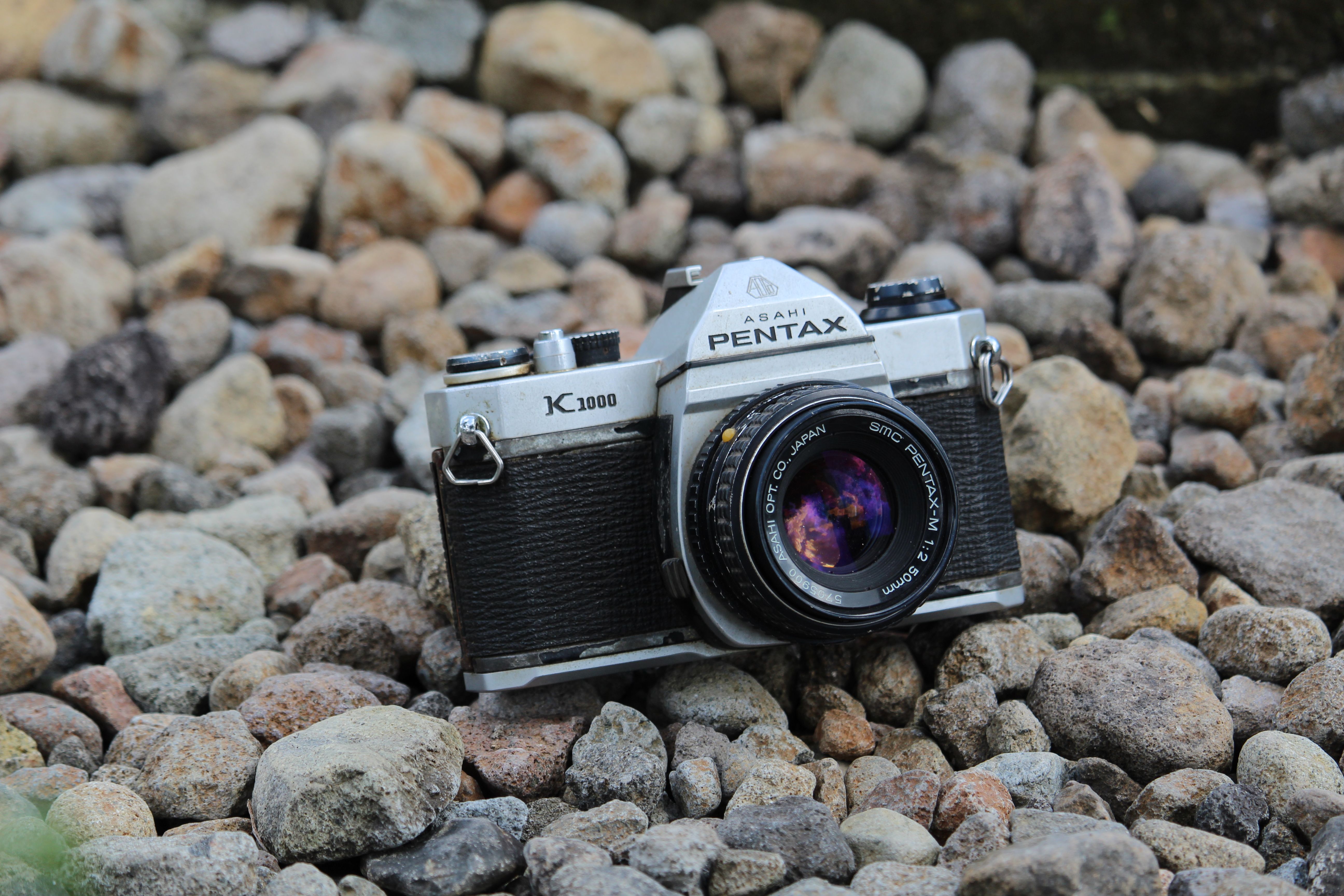 A classic Pentax camera rests on rocky ground in an outdoor setting.