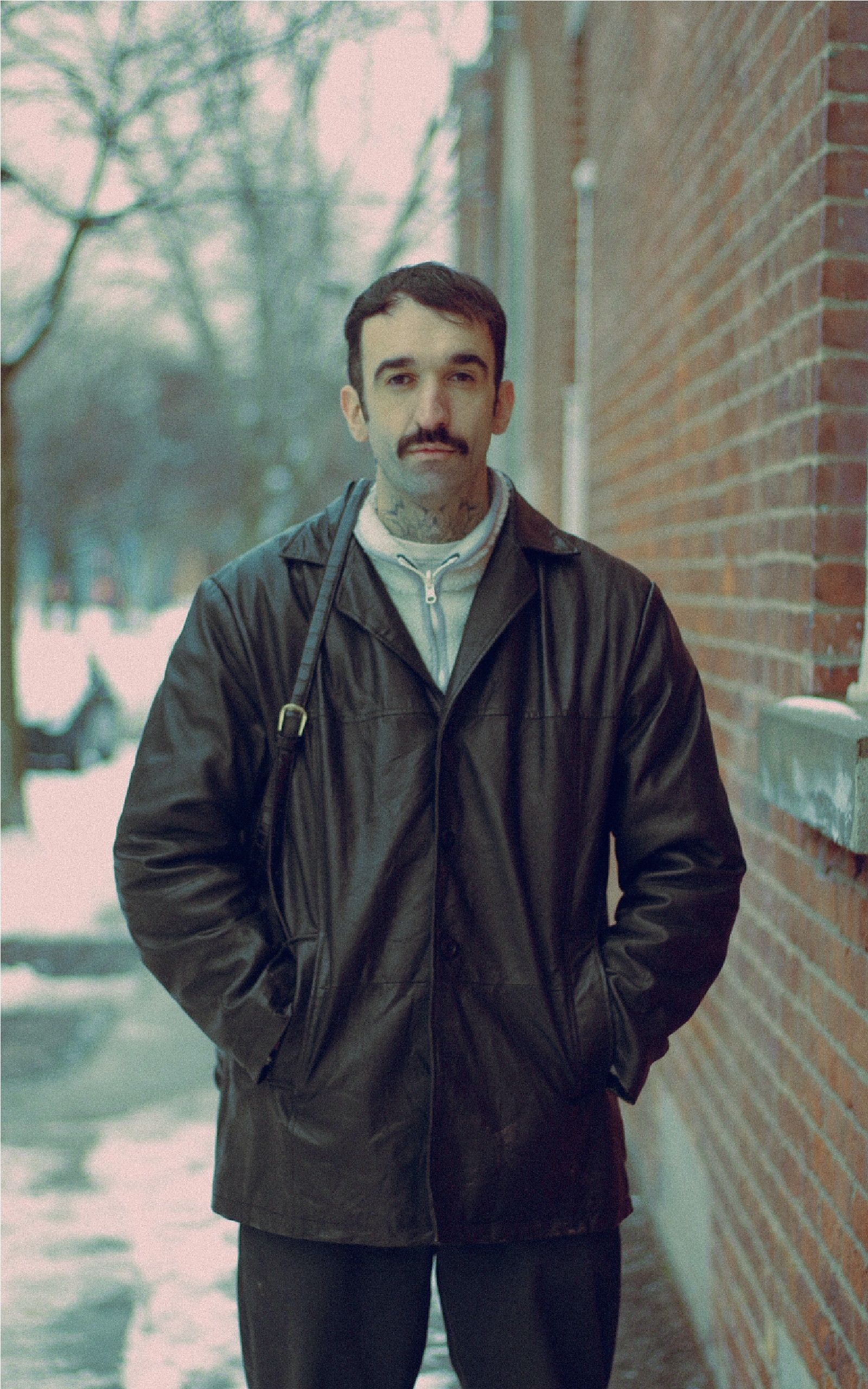 Portrait of a man with a mustache in a leather jacket outdoors by a brick wall in snowy Montreal.