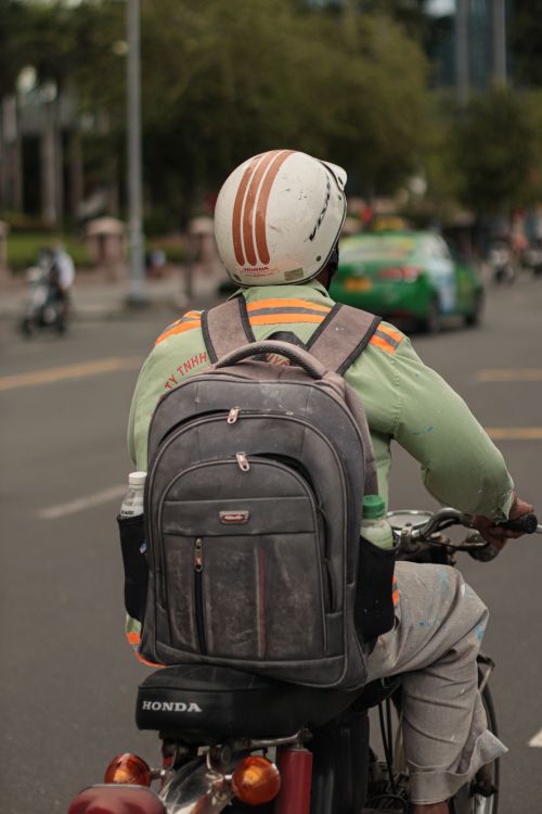 Free A motorcyclist wearing a backpack and helmet rides through a city street, viewed from behind. Stock Photo