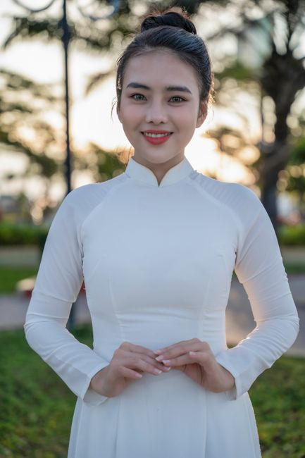 Smiling young woman in a white Ao Dai, standing outdoors with a serene background.