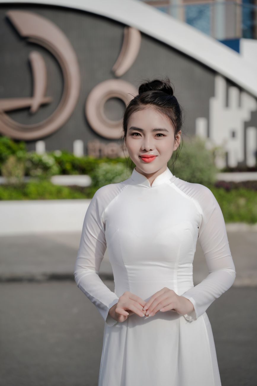 Woman in white Ao Dai poses outdoors with modern architecture backdrop.