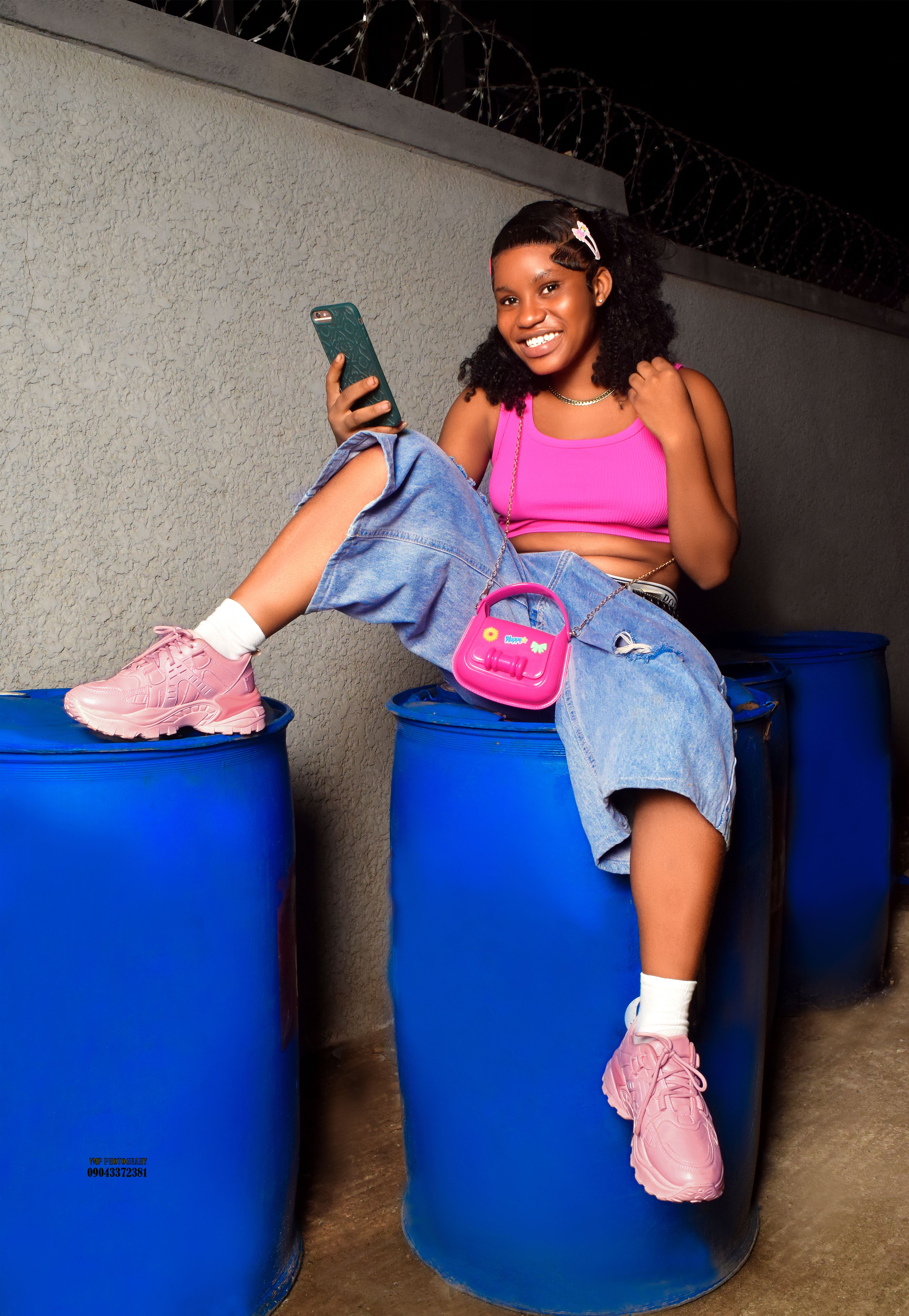 Young woman with smartphone sitting on blue barrels at night, wearing pink and denim.