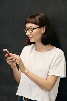 一位戴眼镜的年轻女子在黑板背景前微笑着使用智能手机