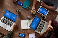 会议中，多元化团队使用技术的顶视图：木桌上摆放着笔记本电脑、平板电脑和智能手机