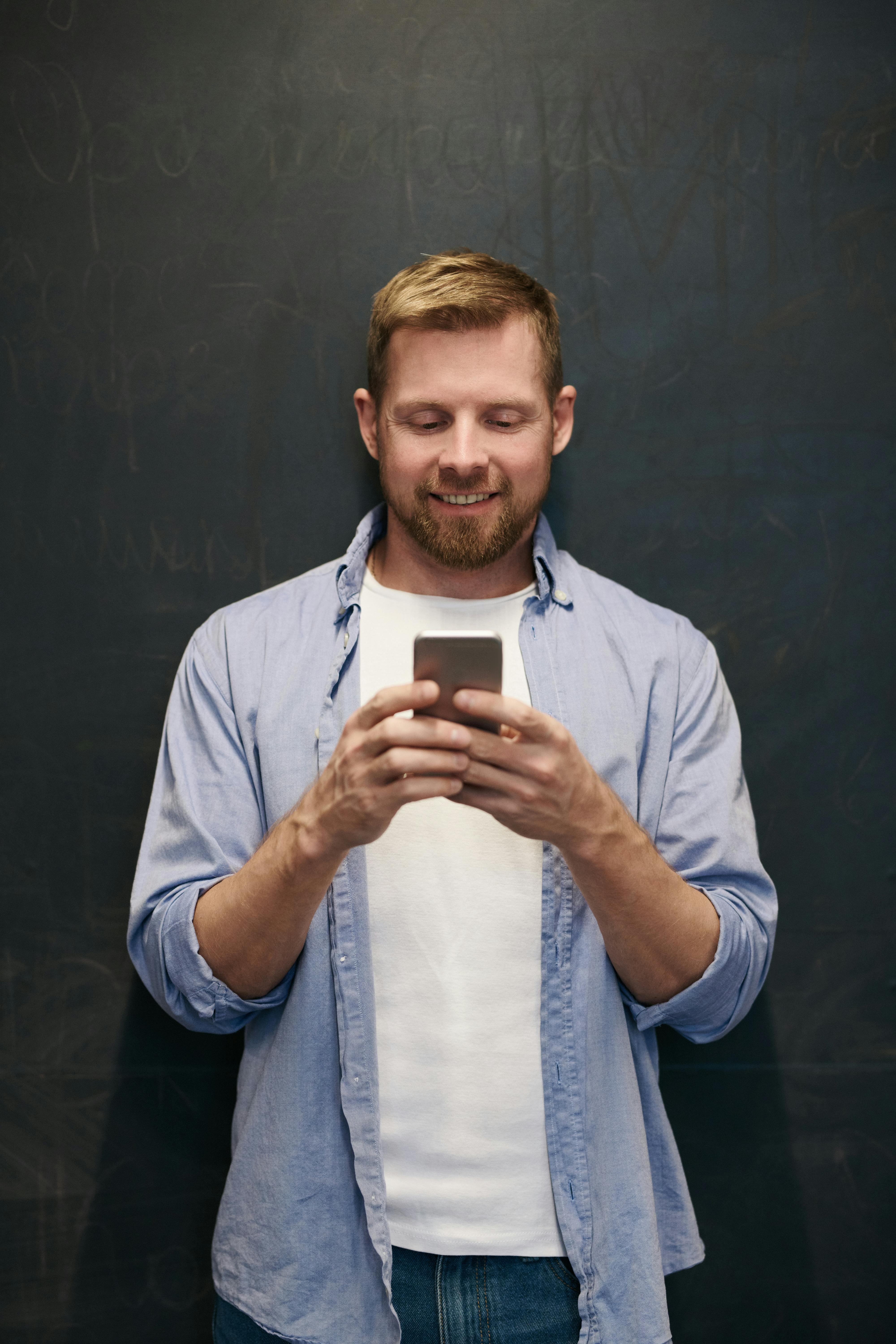 A cheerful man using a smartphone against a chalkboard indoors.