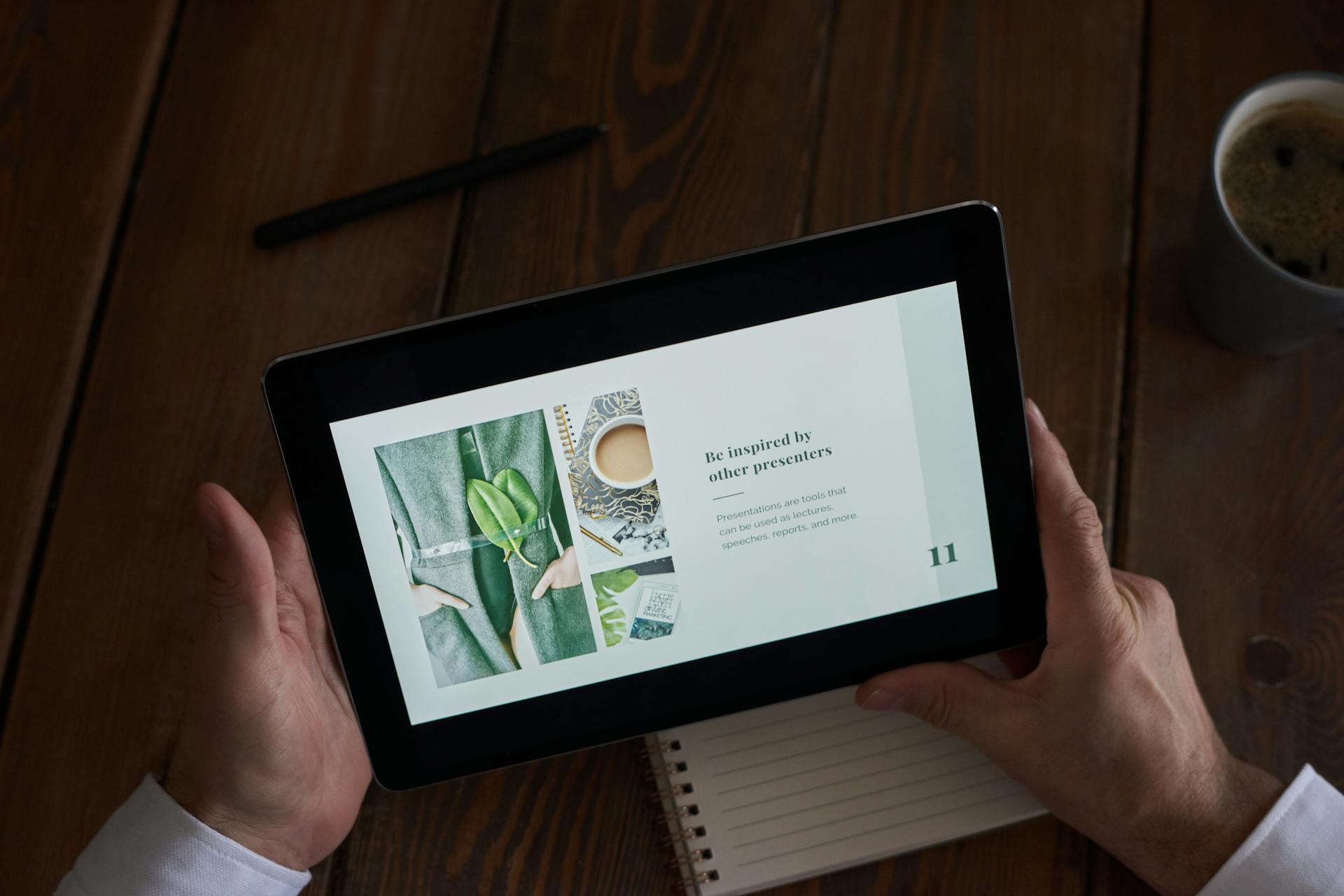 Close-up of hands holding a tablet showing a presentation on a wooden desk.