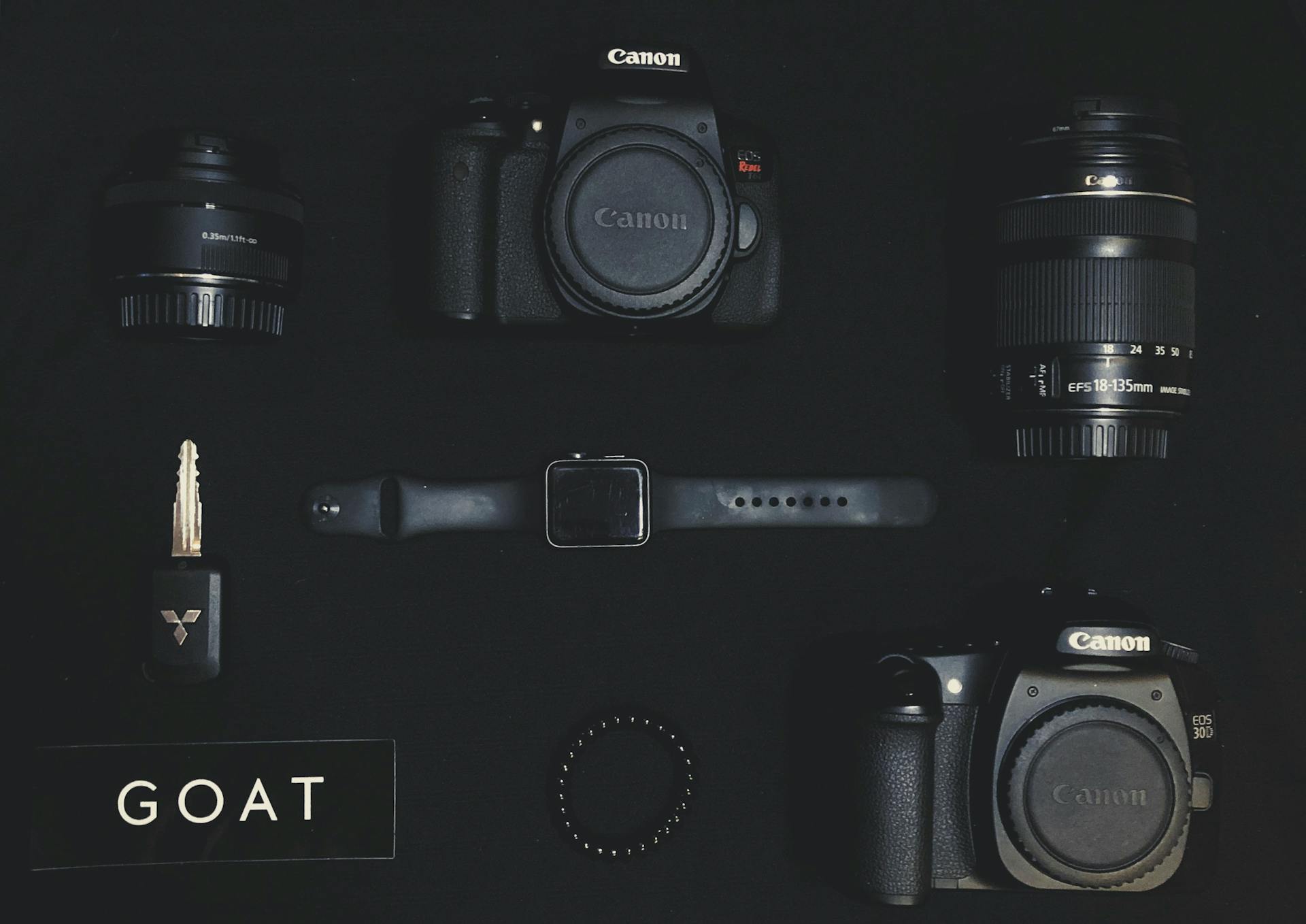 Flat lay of camera gear including Canon, lenses, watch, and key on a dark background.