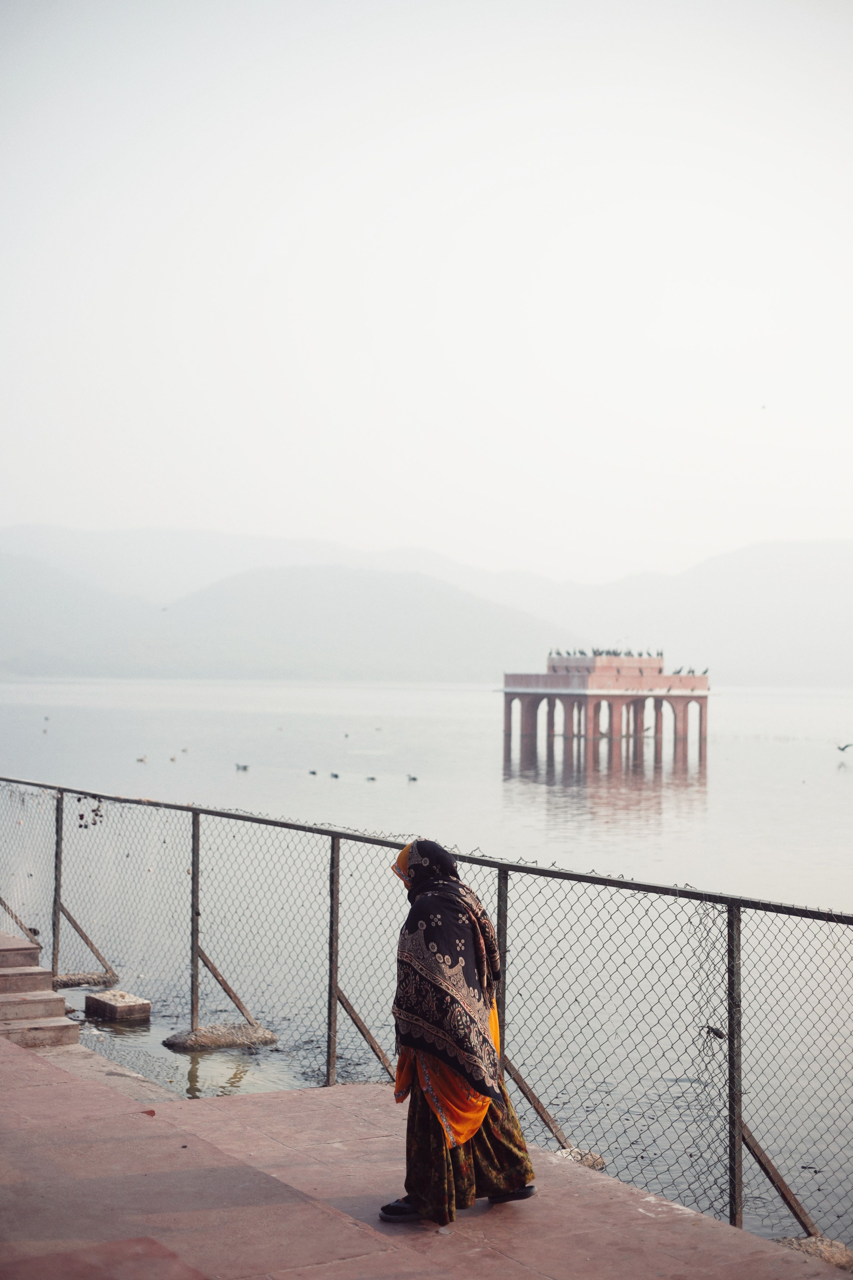 A woman walks along the tranquil Jal Mahal lake in Jaipur, India, surrounded by a peaceful landscape.