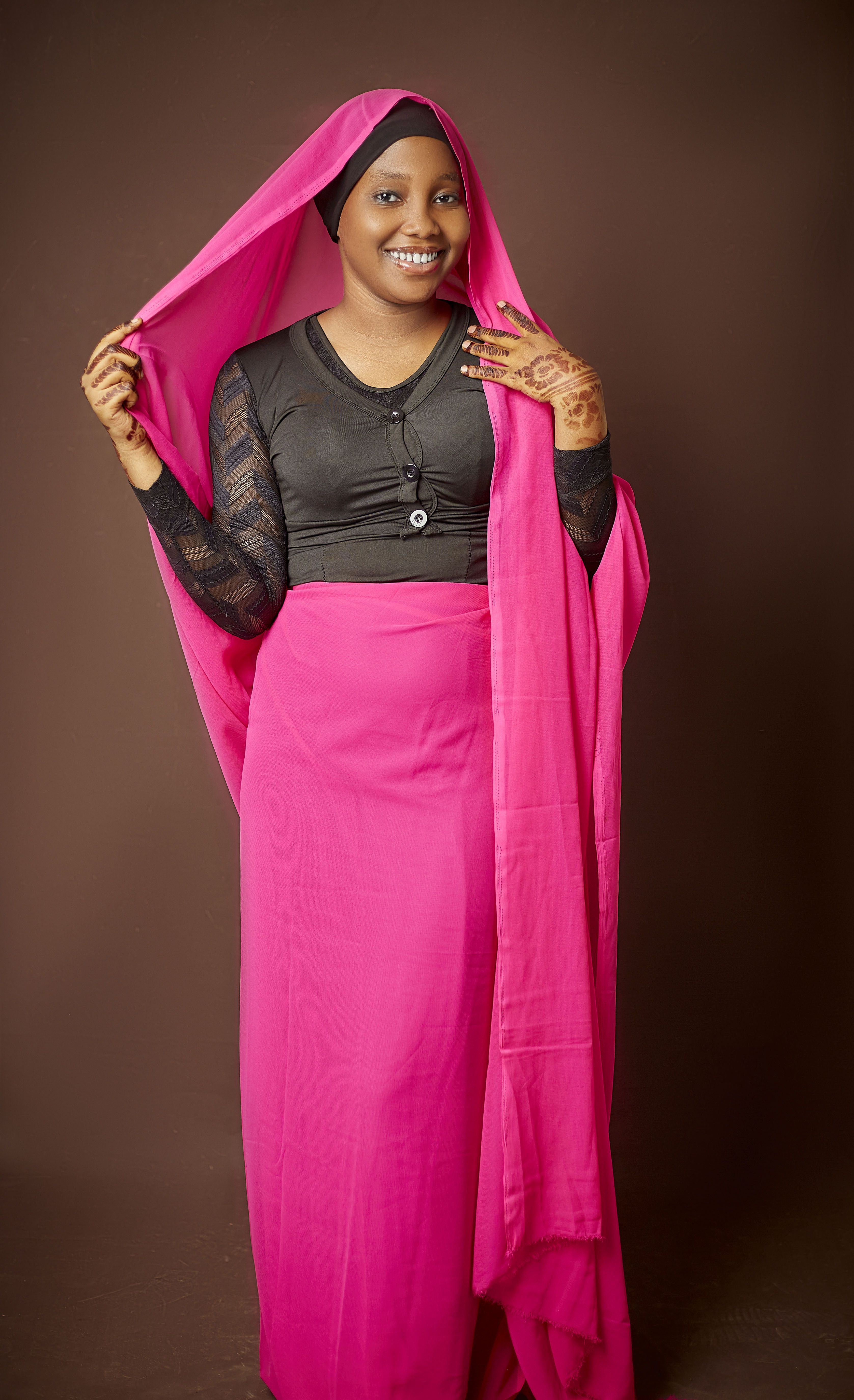 A cheerful woman wearing a vibrant pink scarf and traditional dress stands against a brown background.
