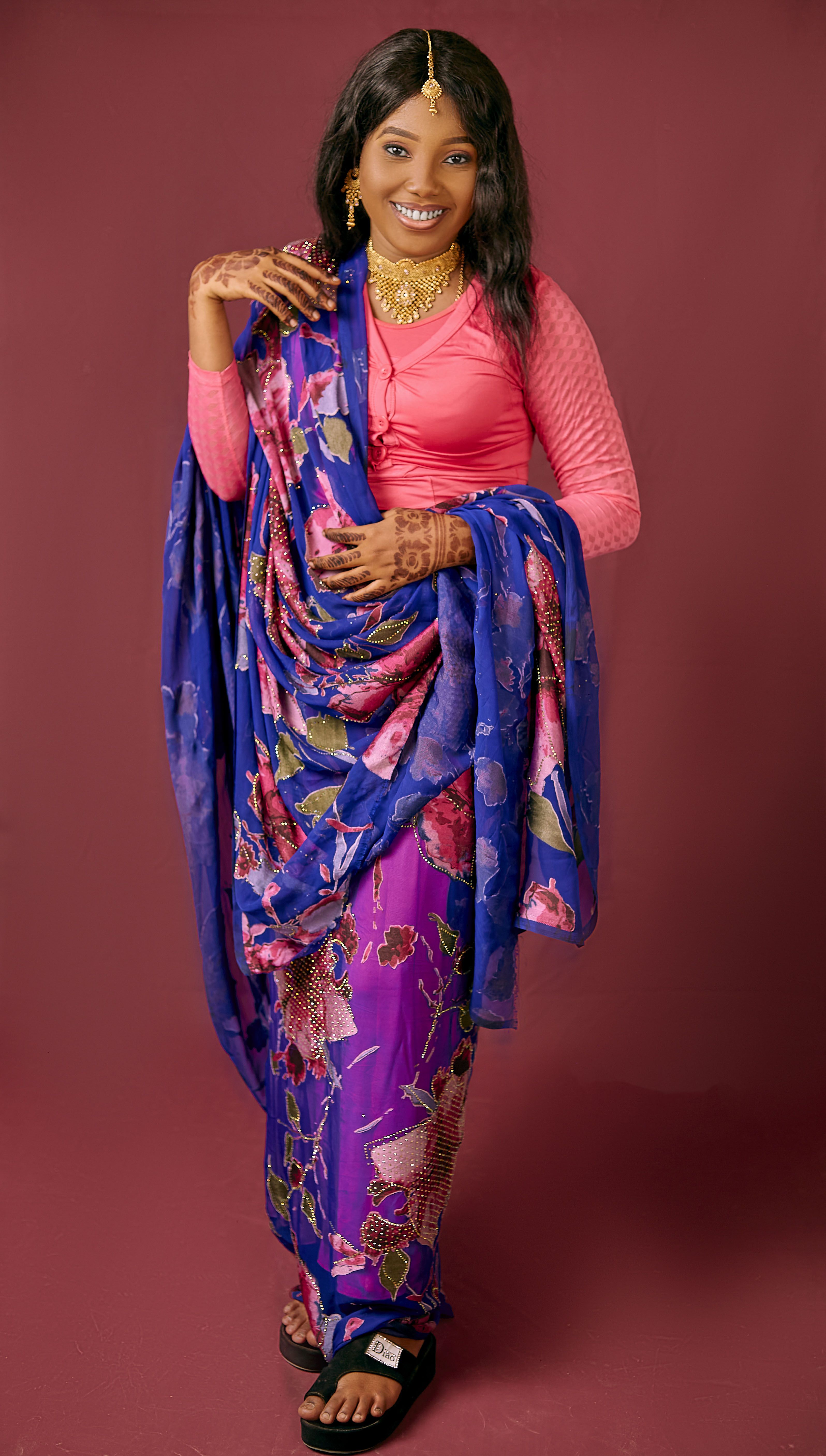 Smiling woman in a traditional vibrant saree poses gracefully against a soft maroon background.