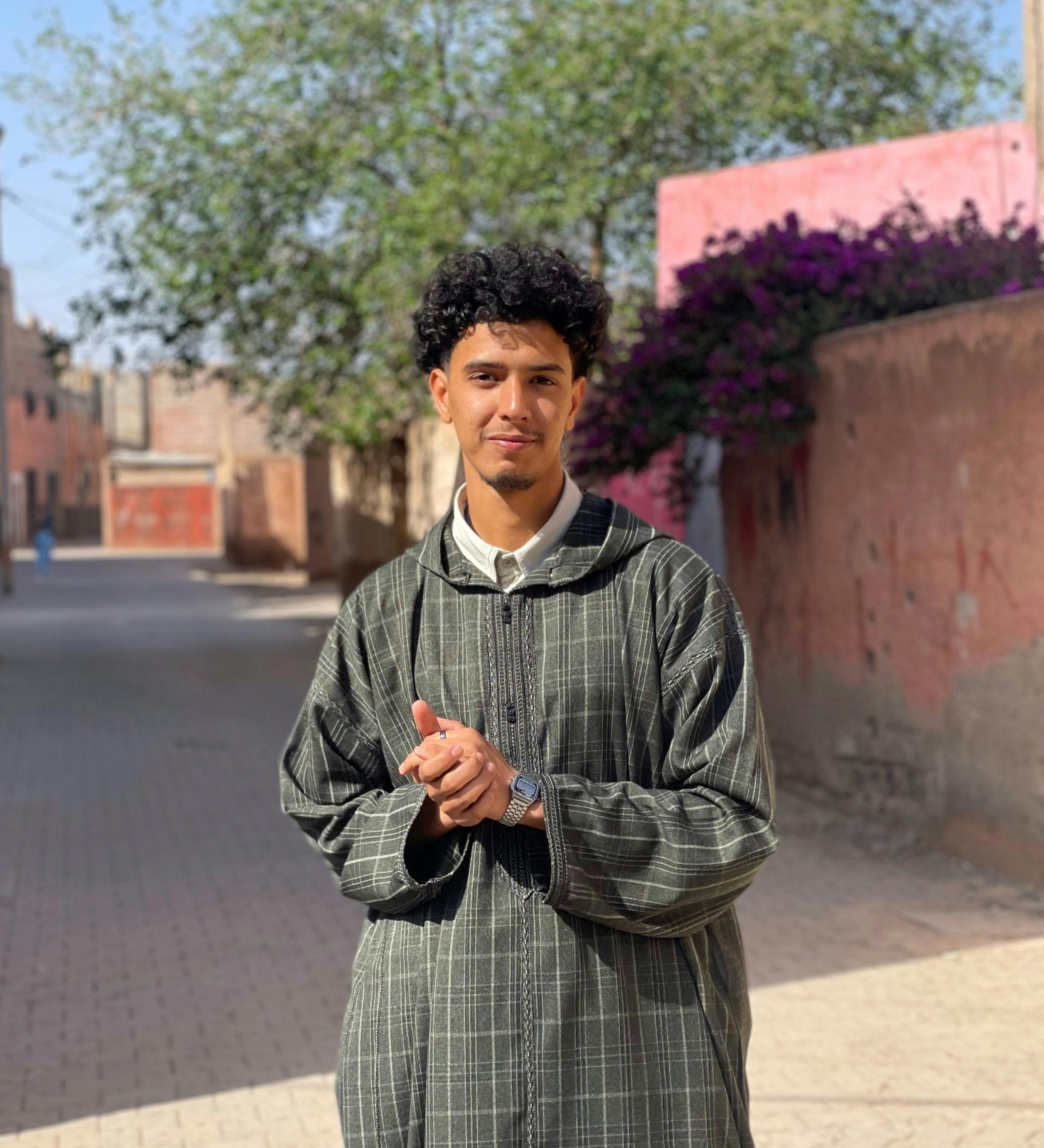 Young man in traditional attire smiling on a sunny street with blooming flowers.