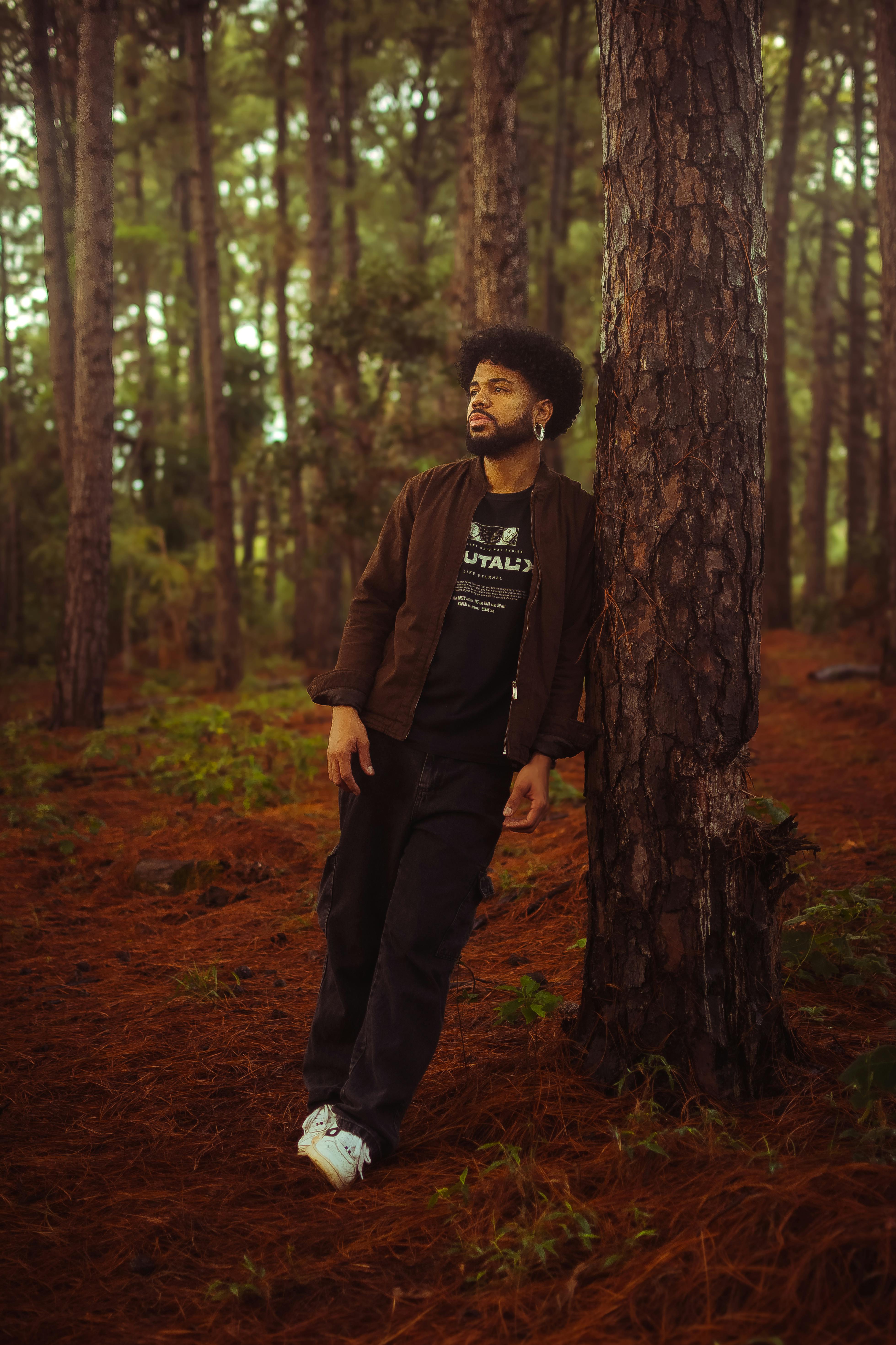 A man leans peacefully against a tree in a serene forest setting, surrounded by tall pine trees.