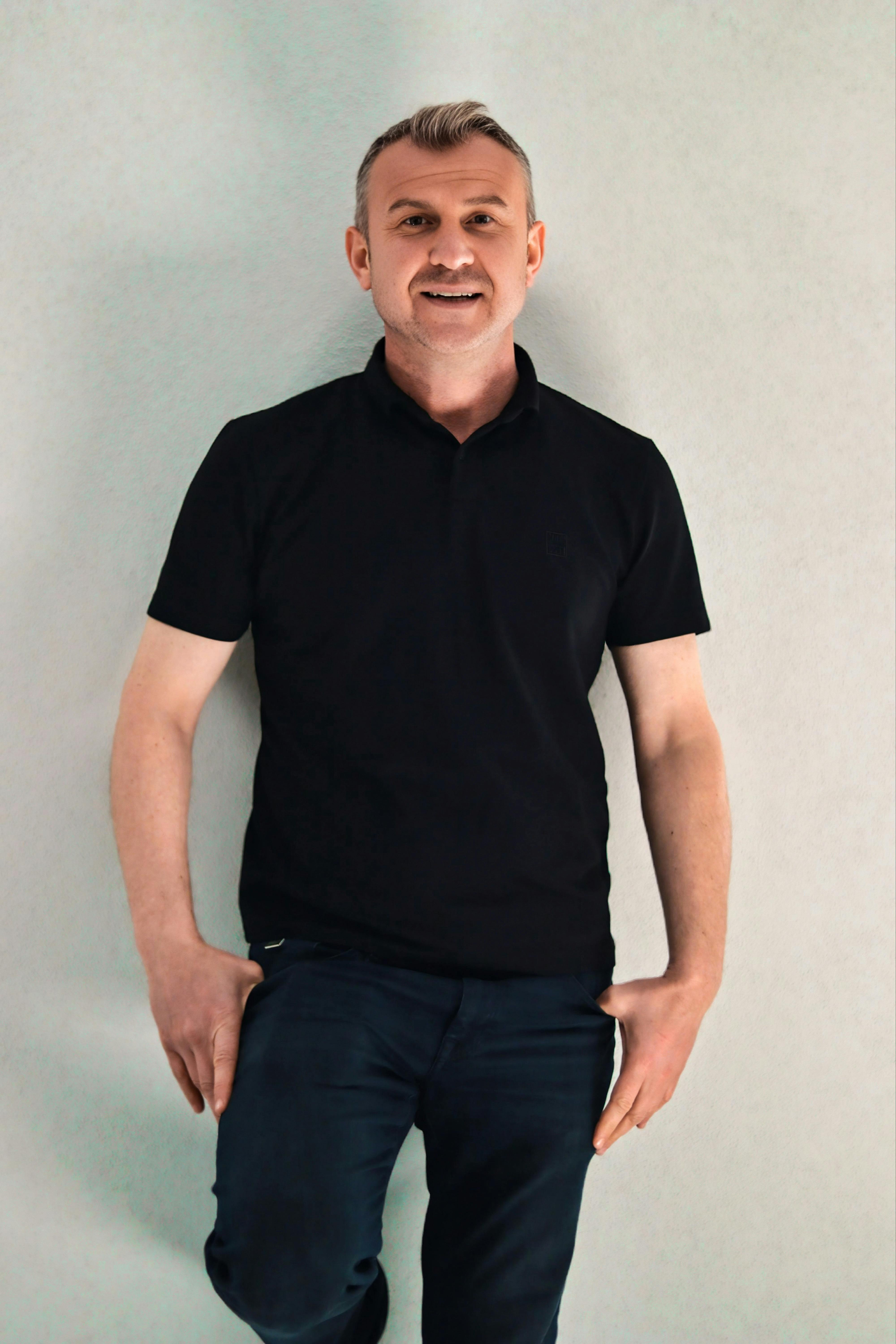 Adult male posing casually with a smile against a plain backdrop, wearing a black shirt and jeans.
