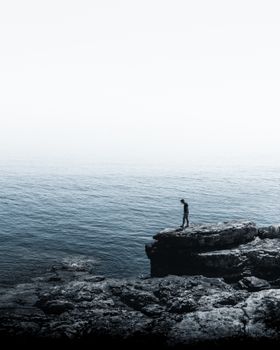 A lone person stands on a rocky cliff overlooking the vast ocean, capturing solitude.