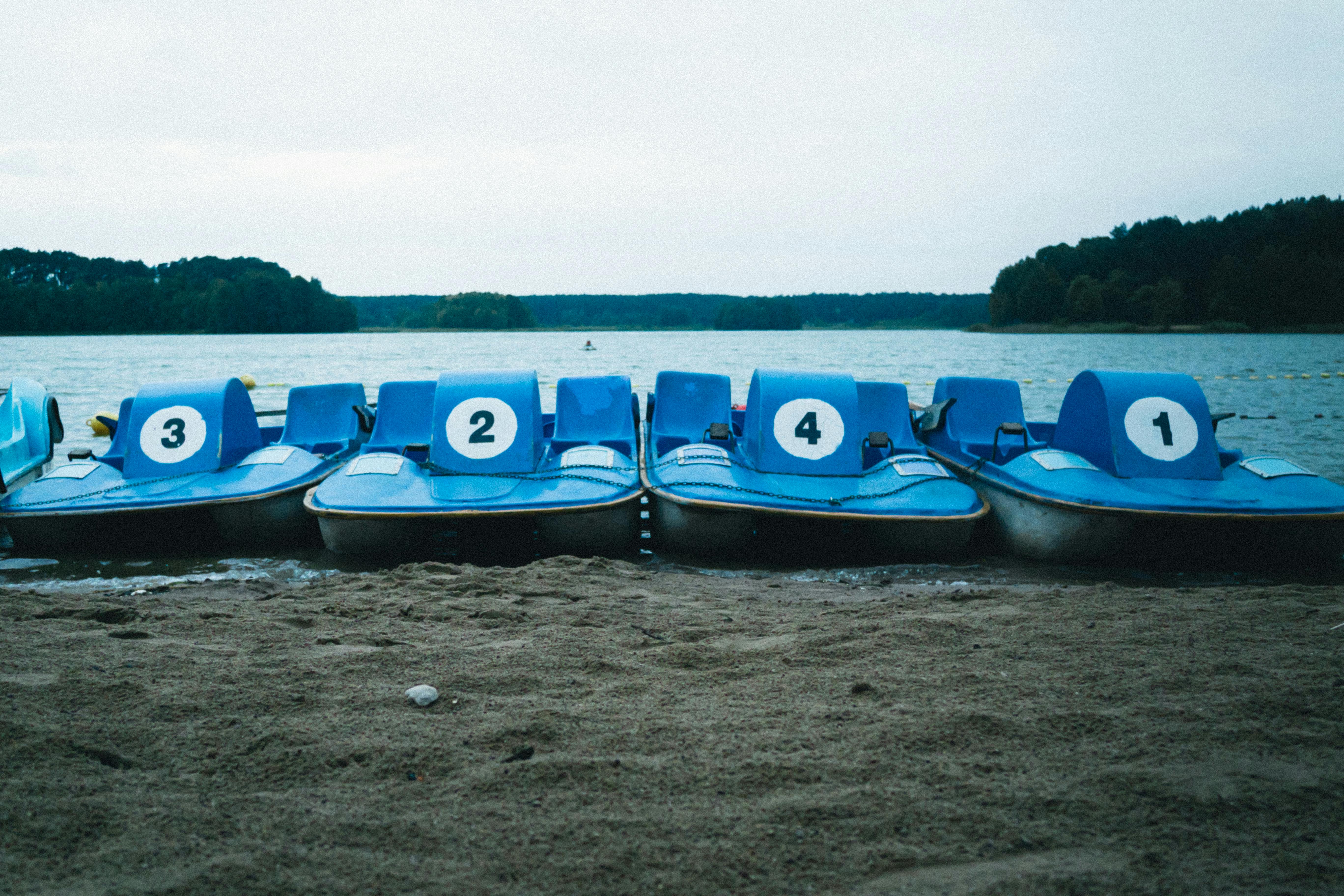 Row of blue pedal boats lined up on a sandy beach by a calm lake under a cloudy sky.
