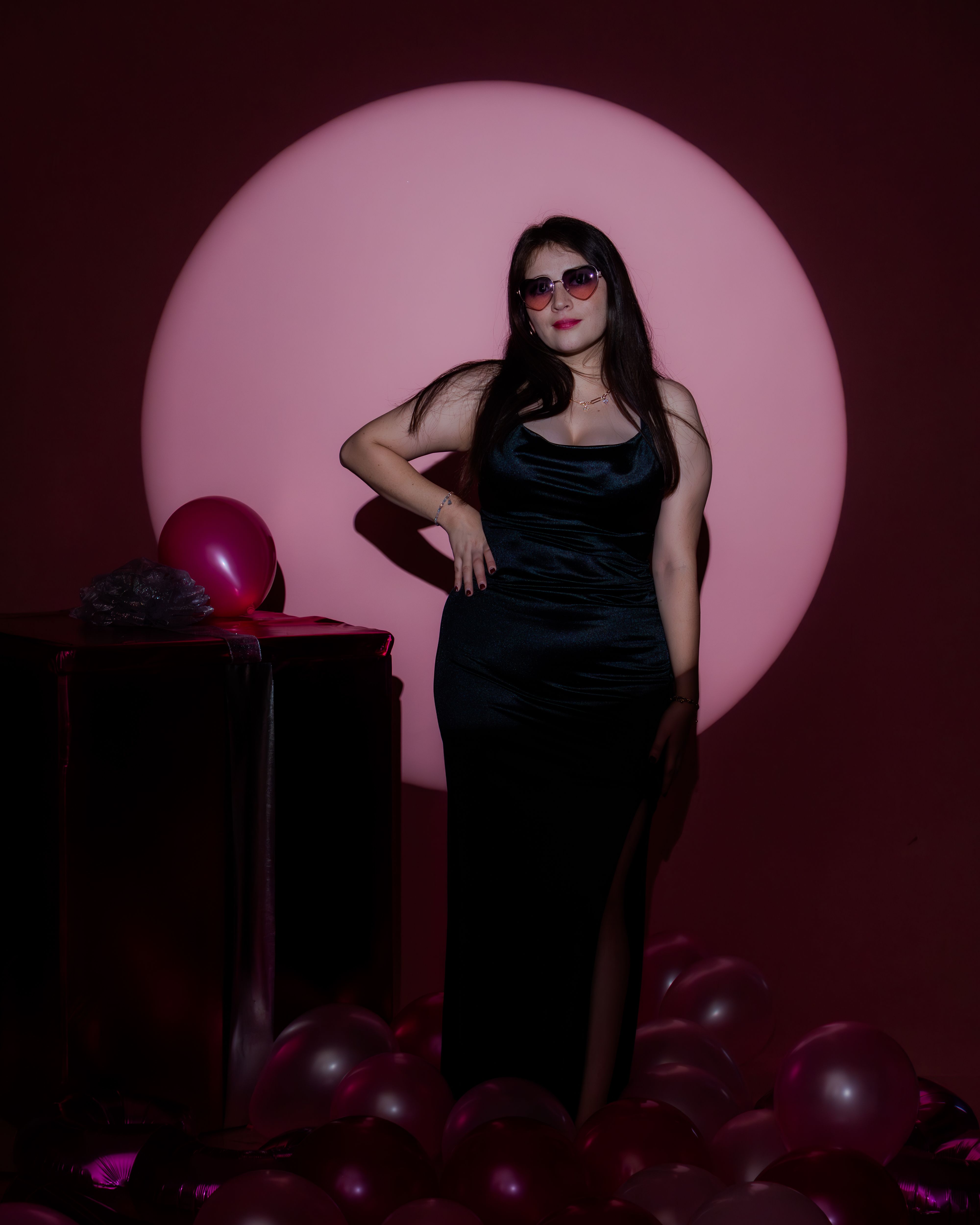 Stylish woman in sunglasses poses amidst a pink backdrop and balloons, exuding elegance and confidence.