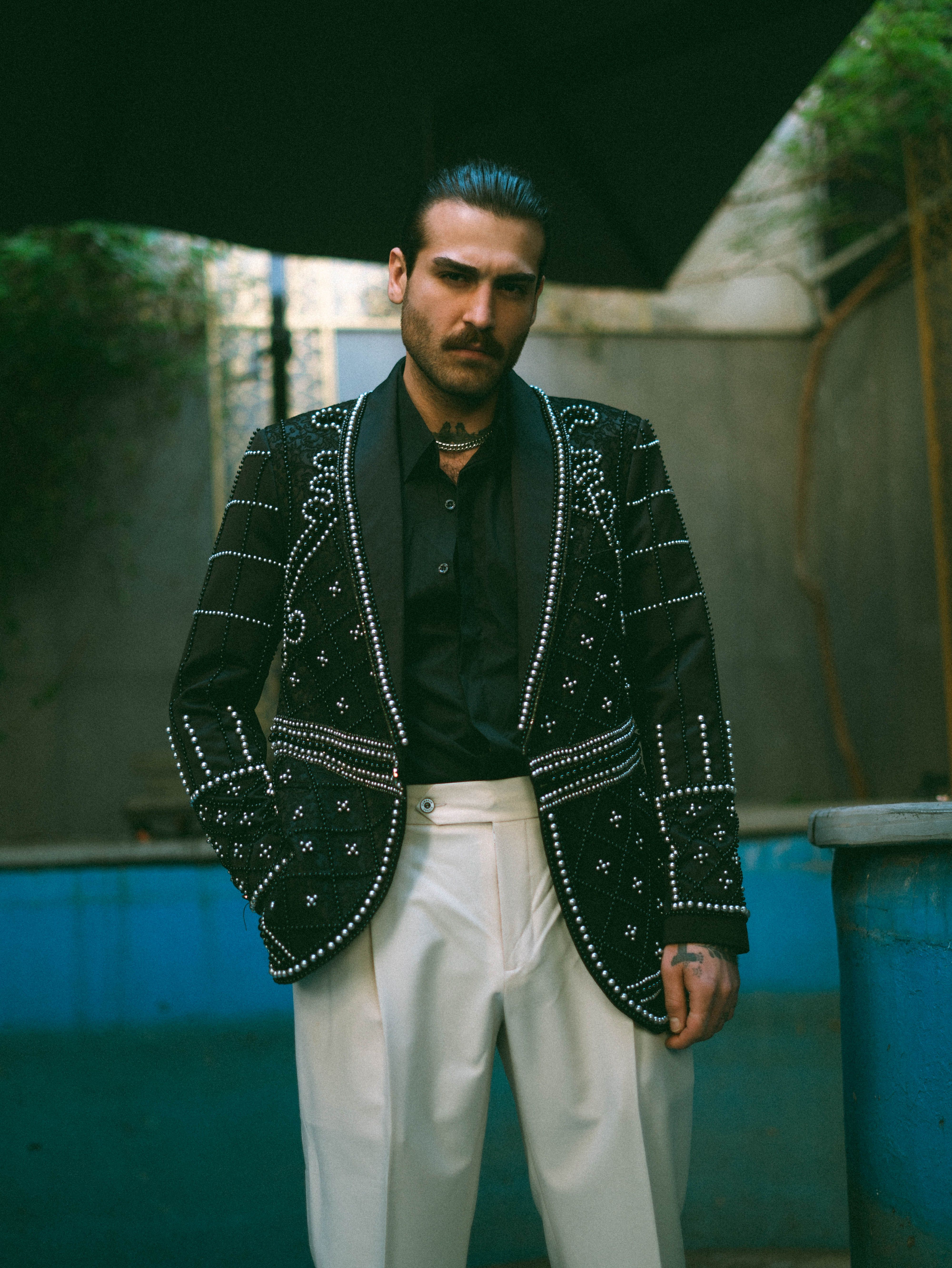 Fashionable man wearing a detailed black jacket and white pants, posing confidently outdoors.