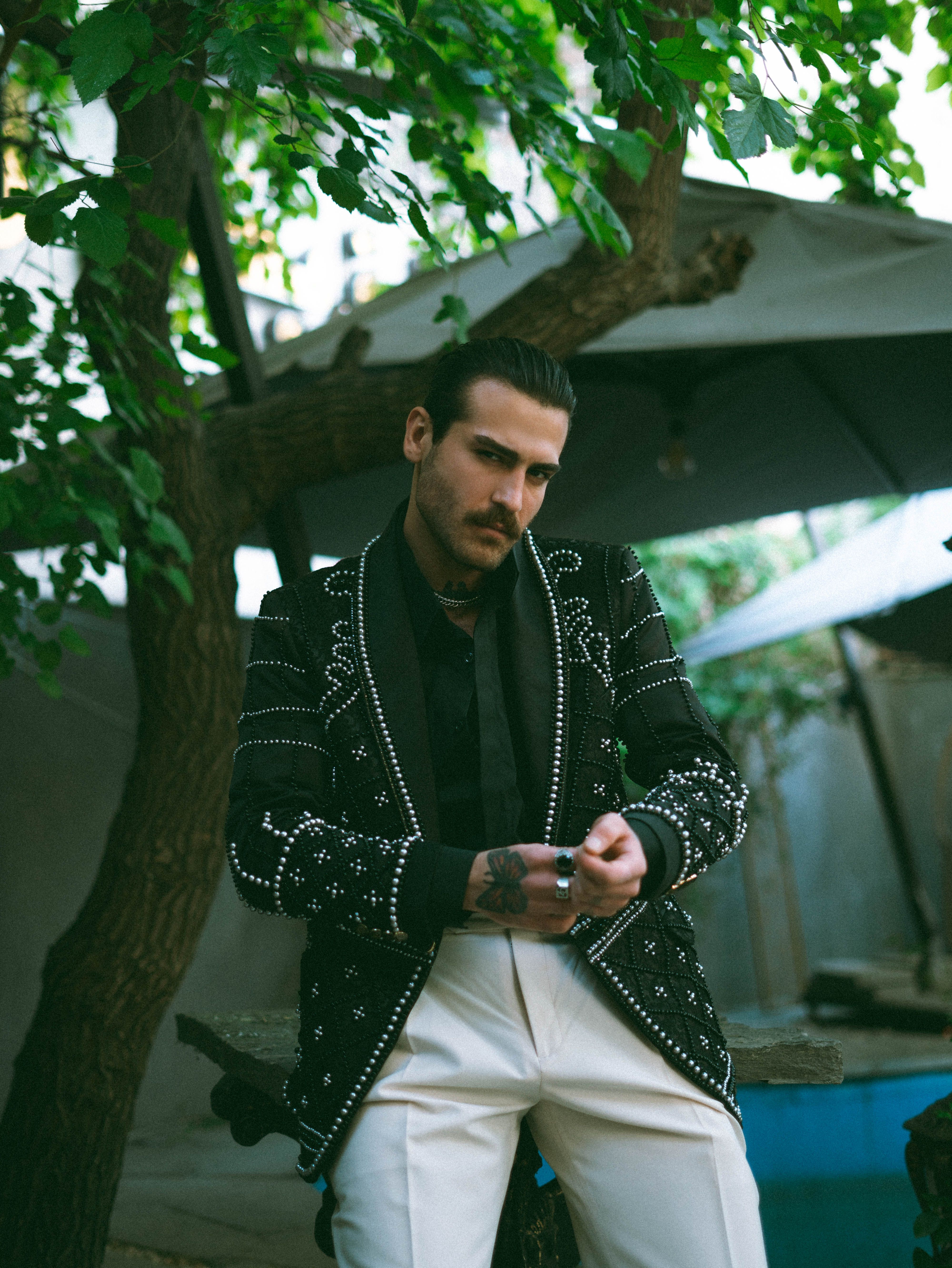 A fashionable man poses in a beaded jacket under lush green trees, exuding style and confidence.