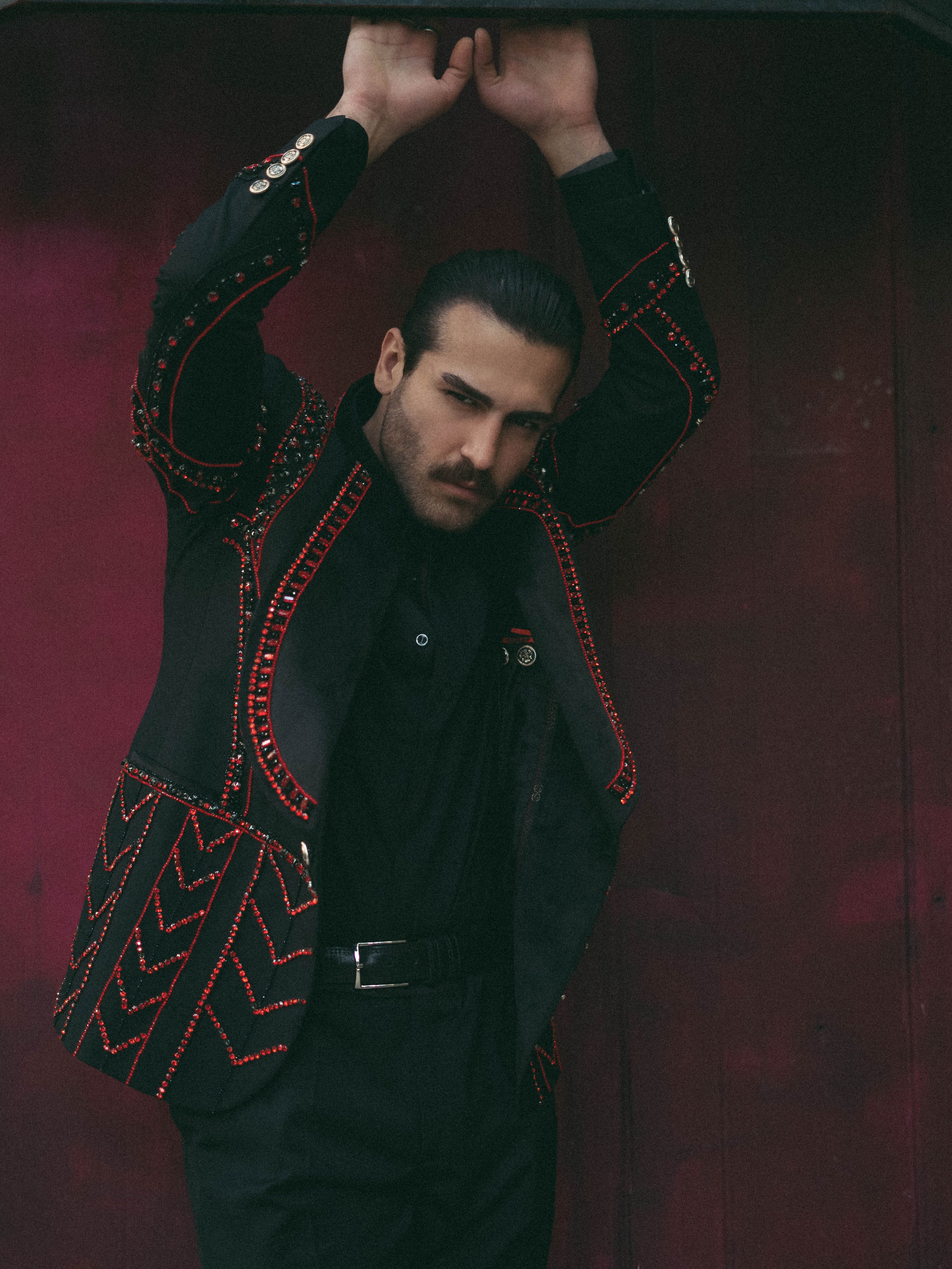 A man wearing an intricately designed jacket poses stylishly indoors with arms raised.