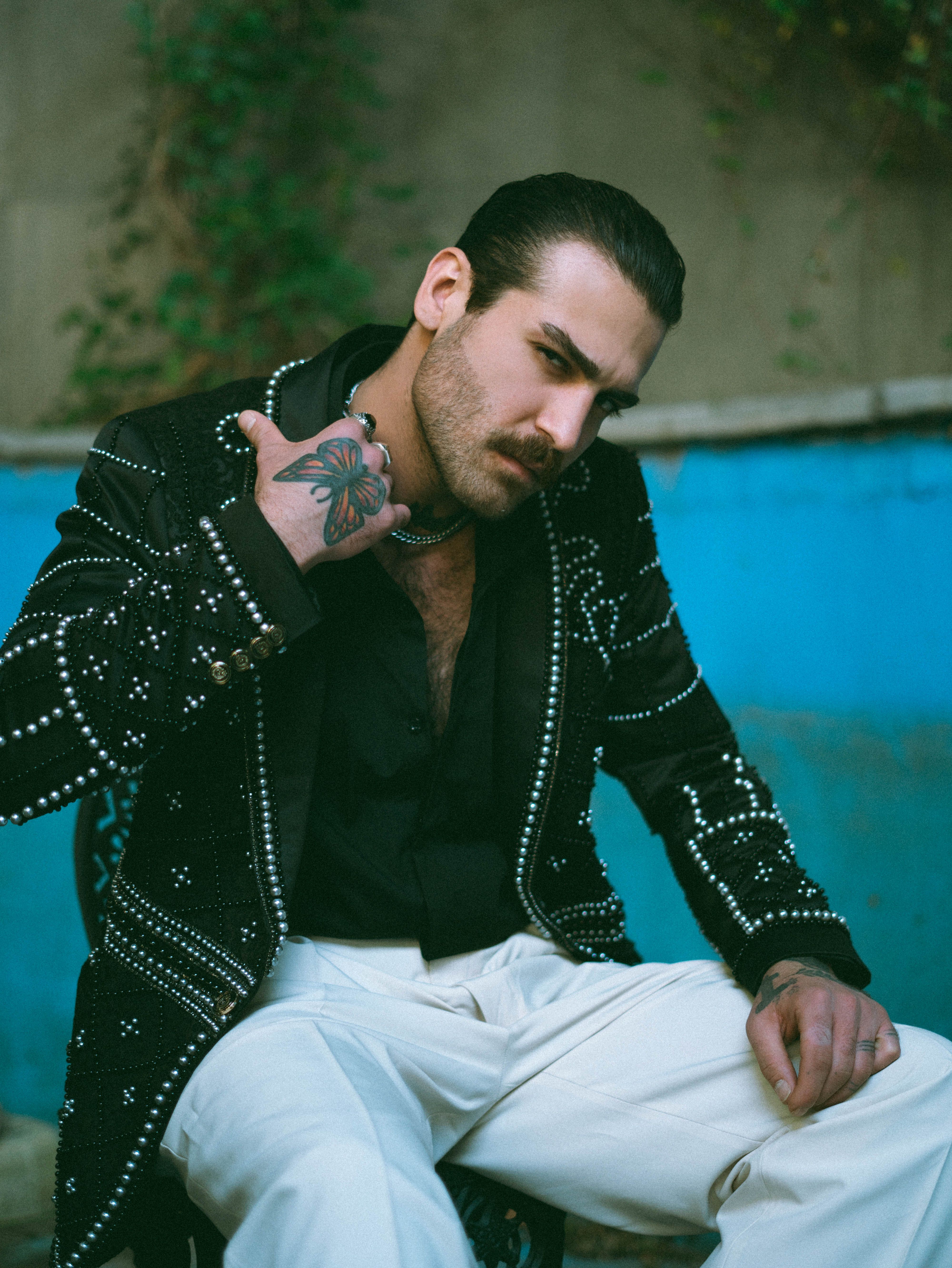 Fashionable man wearing a studded jacket and white pants, sitting outdoors with a confident pose.