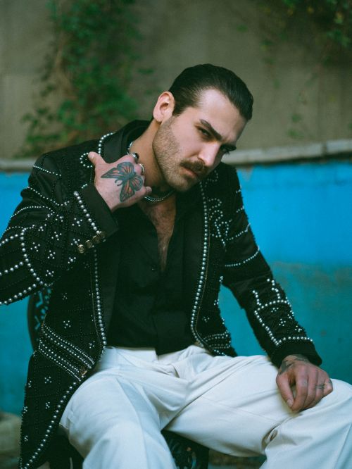 Free Fashionable man wearing a studded jacket and white pants, sitting outdoors with a confident pose. Stock Photo