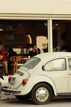 Classic white car parked by a bustling cafe with patrons inside, evoking a retro-modern feel.