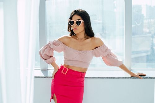 Free Stylish woman wearing heart-shaped sunglasses and pink attire poses by a sunlit window, exuding confidence and modern fashion. Stock Photo