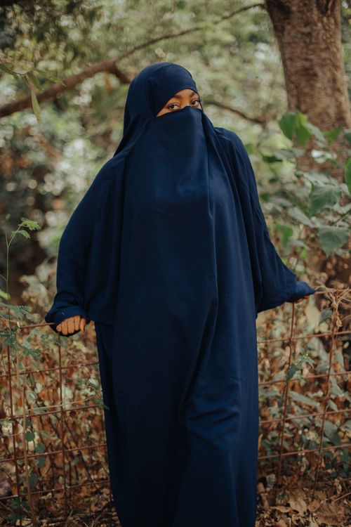 Free A woman in a blue niqab stands outdoors surrounded by lush greenery. Stock Photo