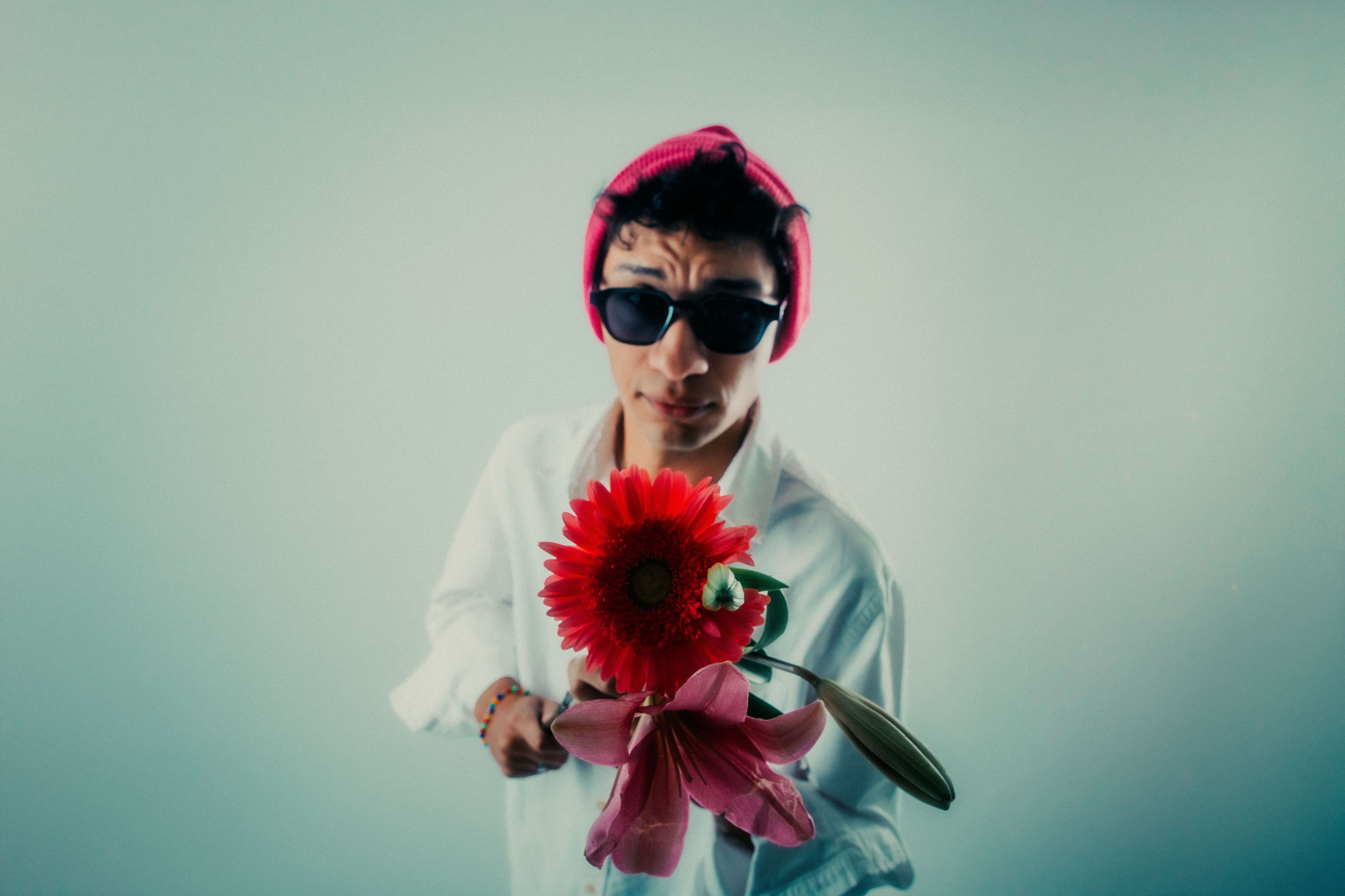 A young man in a pink beanie and sunglasses holds vibrant flowers in a studio setting.
