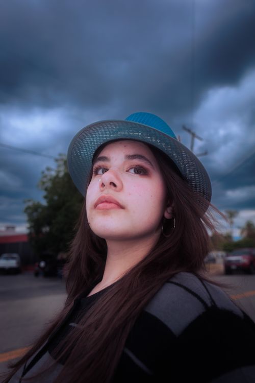 Free A young woman in a blue hat gazes upward against a backdrop of dramatic clouds. Stock Photo