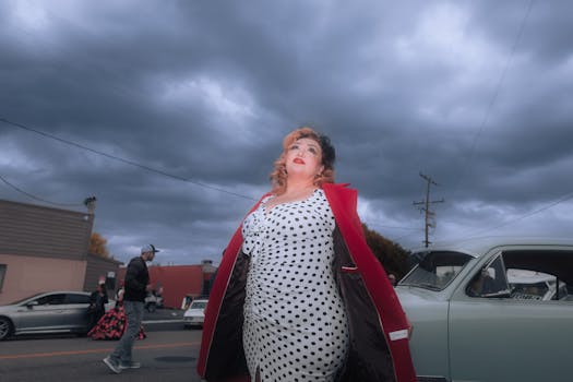 A stylish woman in vintage polka dot dress posing near a classic car under dramatic skies.