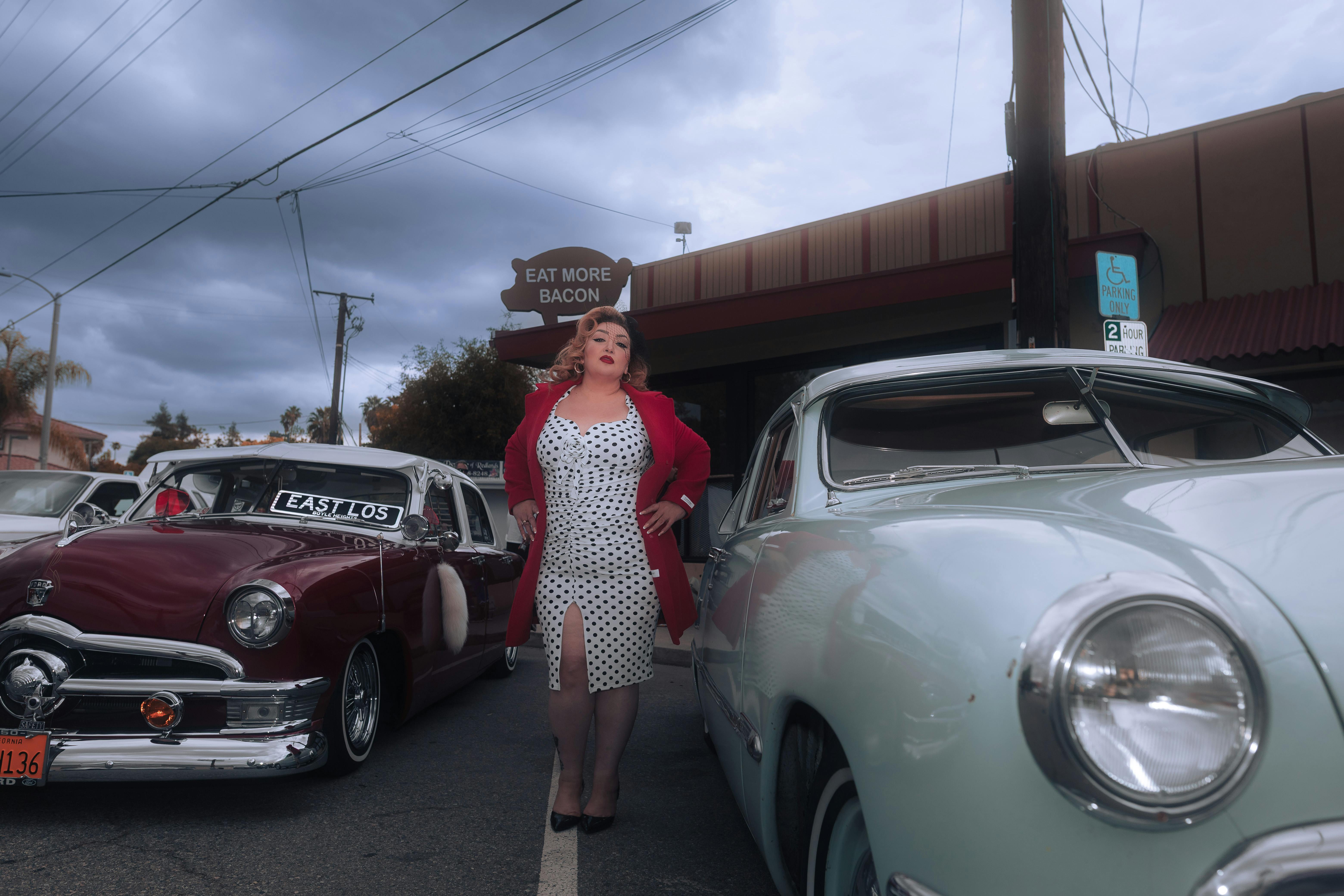 A vintage car show with a retro fashion model posing in front of classic cars on an overcast day.