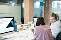 在现代办公环境中，两位发型各异的女性正使用台式电脑进行合作